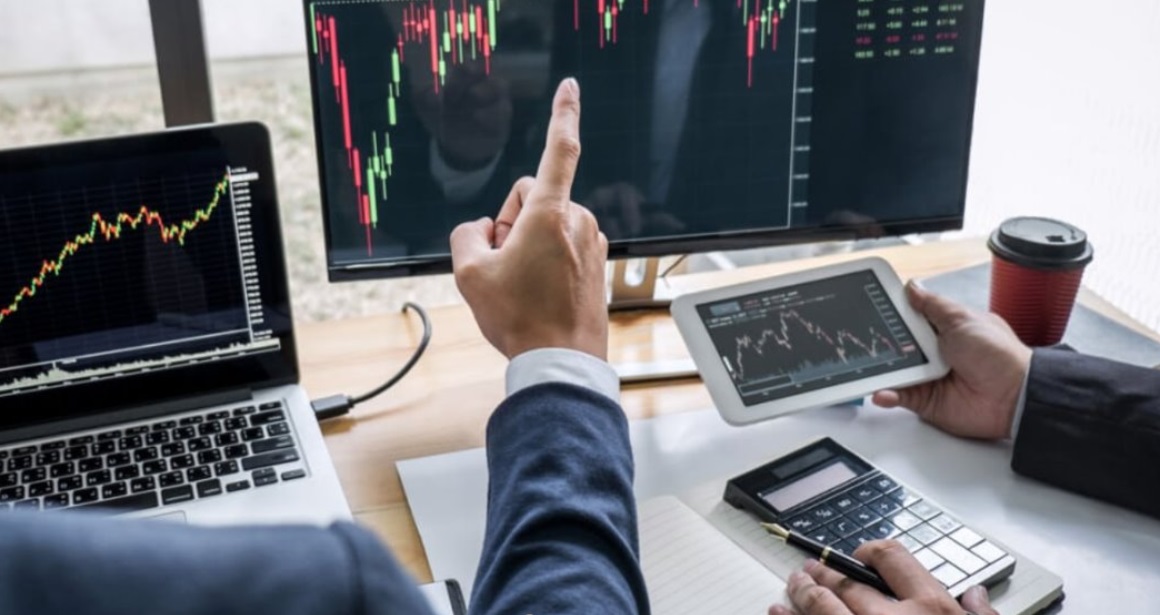 Two people are discussing trades in front of a computer displaying a trading chart.