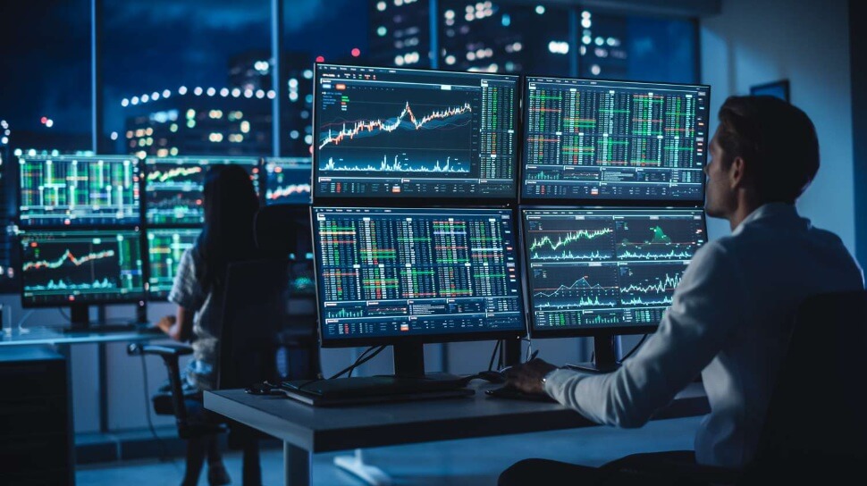 A man and a woman are studying financial trading charts on monitors.