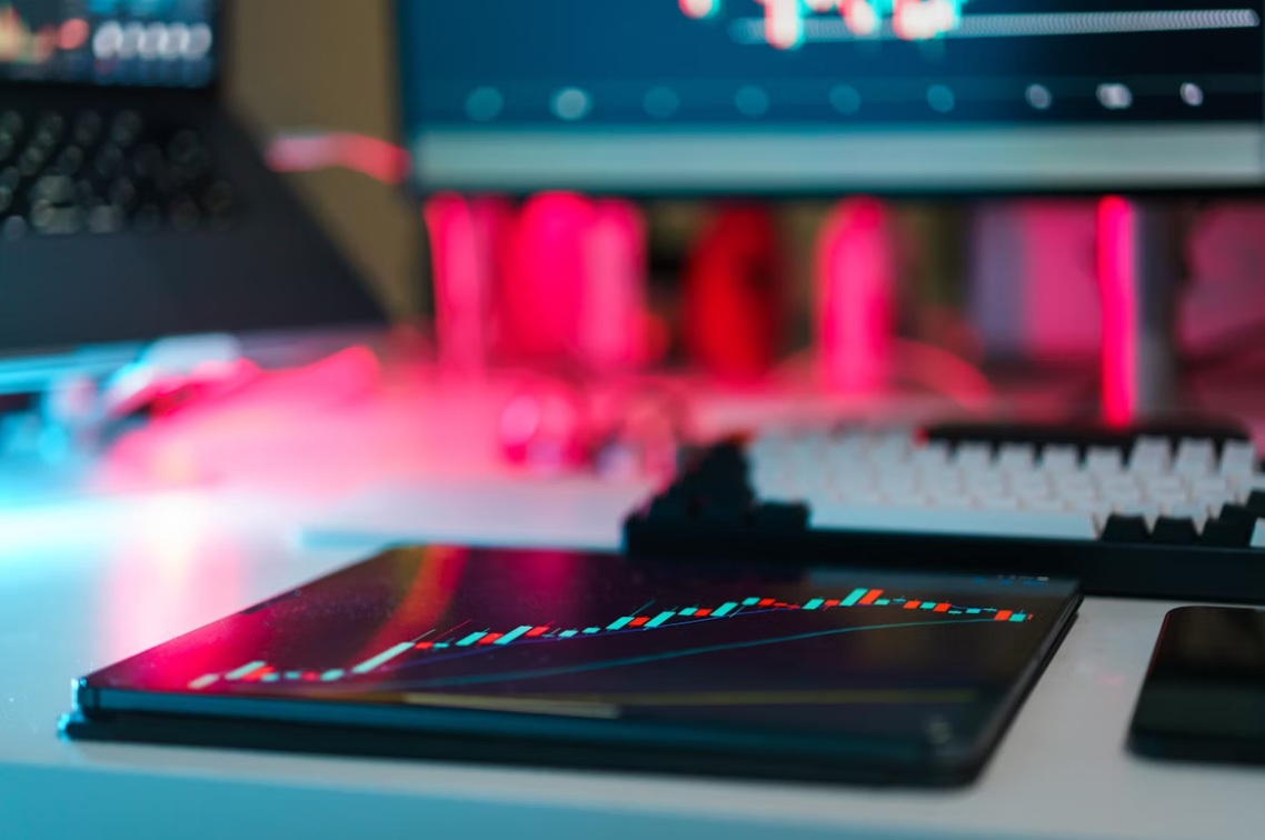 A tablet shows a candlestick chart, while a desktop computer appears blurred in the background.