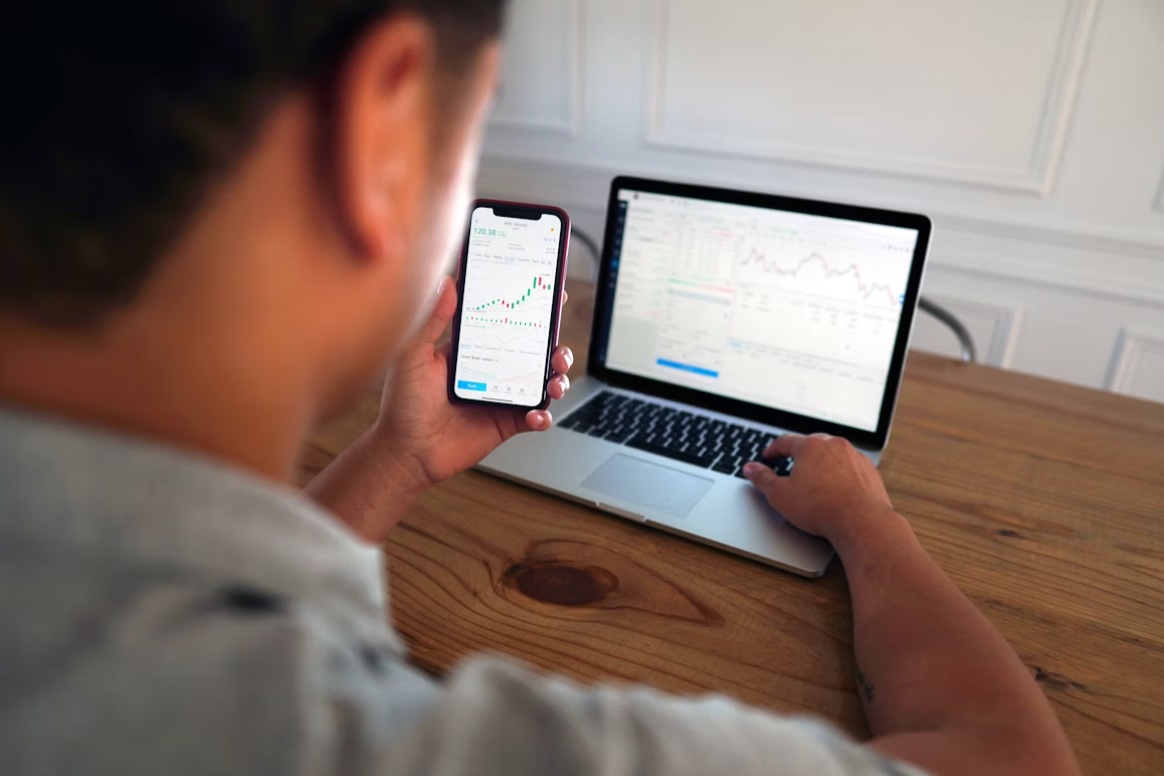 A man is holding a smartphone while analyzing trading charts on his laptop.